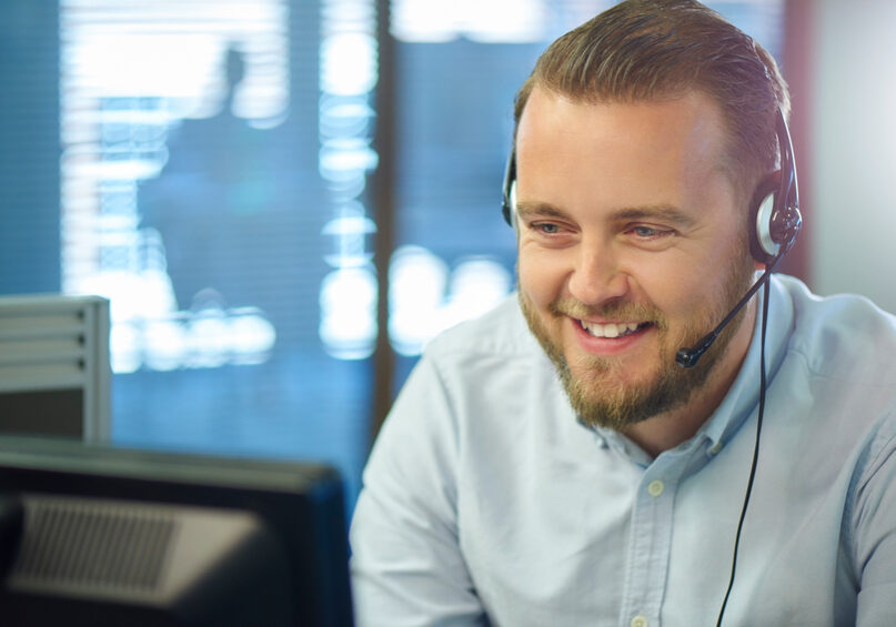 a bearded call centre representative is happily chatting on a headset to a customer . He is sitting in an open plan call centre , with a manager's office in the background with an unrecognisable meeting taking place .